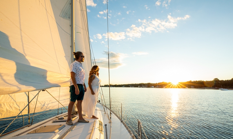 a couple stands on a sailboat looking at the setting sun